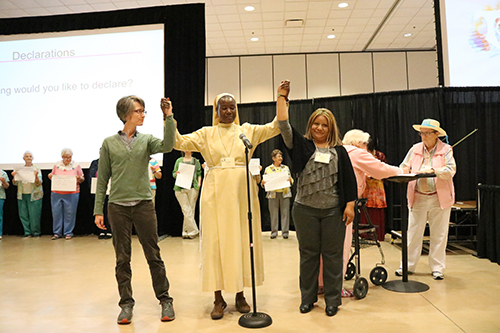 Franciscan Sister of Perpetual Adoration Kristin Peters, Little Sister of St. Francis Sarah Nakyesa and honored guest Reyna Mendez hold raised hands proclaiming A Revolution of Goodness