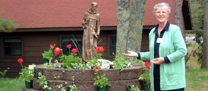 Sister jolynn brehm stands in front of a bed of red flowers and a statue of st francis of assisi