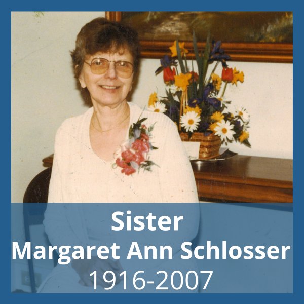 Picture of a woman seated with short brown hair, glasses and a dark pink corsage wearing white in front of a painting, brightly-colored flower bouquet in a basket and a white wall