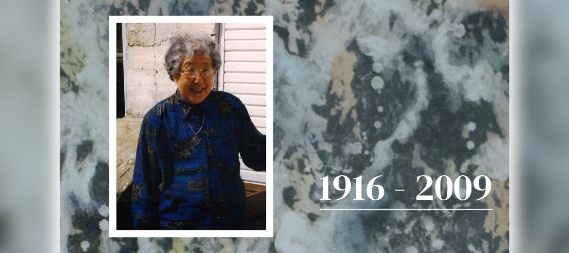 Photo of woman of Asian decent with short gray hair, glasses and silver medallion necklace wearing dark blue and gold shirt in front of a wall of white stone and siding over a muted watercolor abstract painting