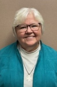Woman with short white hair and glasses wearing a white turtleneck, teal jacket and silver medallion against a light brown background