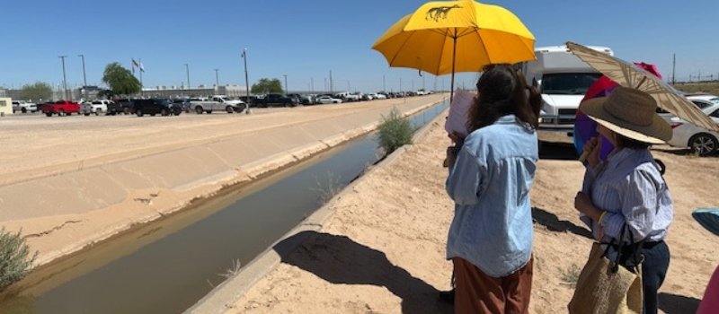 people holding umbrellas to protect them from the sun stand facing the Eloy Detention Center during a prayer vigil