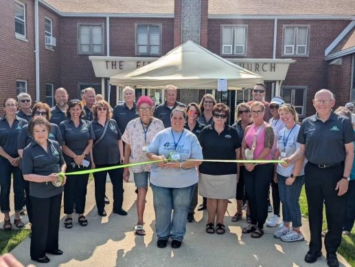 Image of a ribbon cutting event with a large group of people stand before a two-story brick building with two people stretching a neon green ribbon in front of another holding a pair of scissors