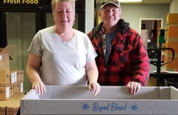 Woman with short blonde hair and white t-shirt with man in baseball cap and red and black-checked flannel shirt touching a wooden grey cart reading Beyond Blessed in blue paint with boxes and a sign reading Fresh Food in the background