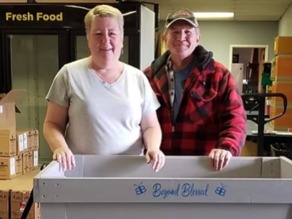 Woman with short blonde hair and white t-shirt with man in baseball cap and red and black-checked flannel shirt touching a wooden grey cart reading Beyond Blessed in blue paint with boxes and a sign reading Fresh Food in the background