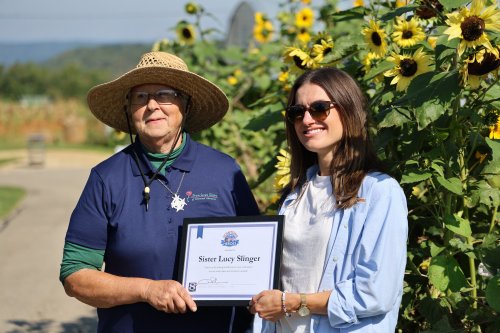 Two women standing outside before tall sunflowers, one wearing a sun hat and glasses, the other wearing sunglasses, both holding an award.