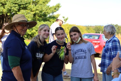 Woman wearing straw hat and glasses standing with three female students holding produce.