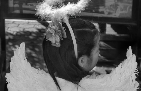 Black and white photo shot from behind of a girl with black hair, wearing a halo head band and feathered wings