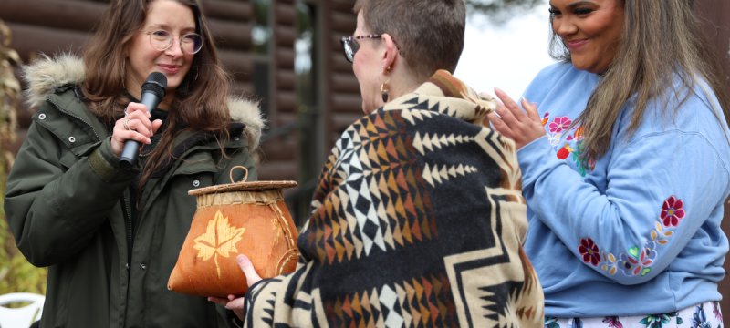 Woman with long brown hair wearing glasses and a fur trimmed jacket with a microphone with a second woman with short, dark hair wearing a white, brown and black native-print shawl holding a basket and a third woman with long brown hair wearing a colorful blue outfit