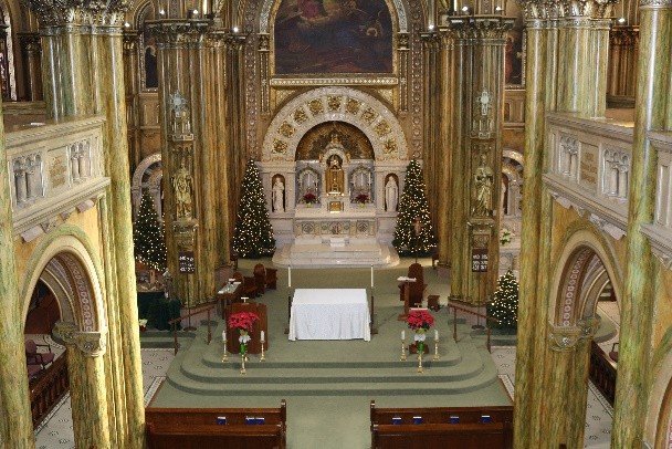 An image of Mary of the Angels Chapel adorned with golden arches and columns above an altar featuring lit Christmas trees, red poinsettias, a table clothed in white and monstrance.