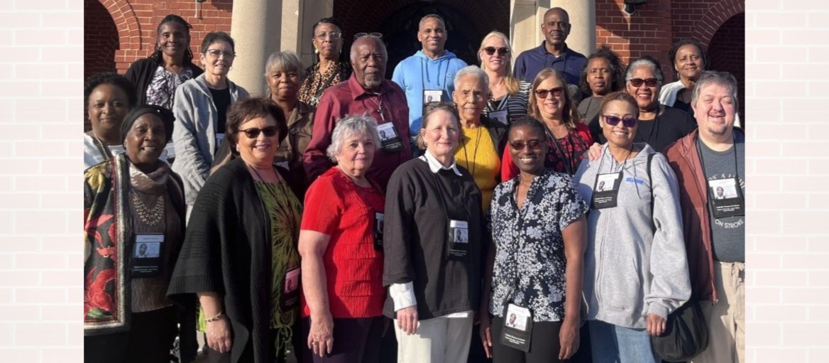 Group of 21 people standing outside in the sun in front of a brick building with two cement columns surrounding a door.