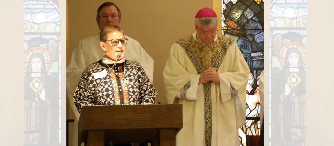 Woman with short hair and glasses wearing a white turtle neck and black and white sweater speaks at a podium in front of a bishop with pink cap and another clergy in white.